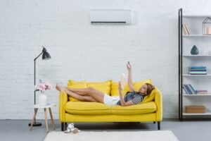 attractive young woman reading book on sofa and pointing at air conditioner with remote control