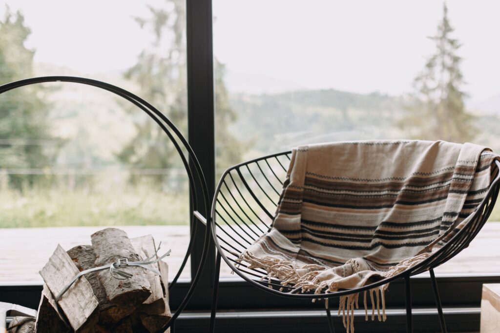 Modern chair with cozy blanket and firewood on metal stand at window view on mountain in cabin