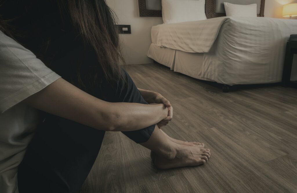 Depressed and stressed woman sitting on vinyl tiles floor near bed in hotel bedroom. Sad woman sit