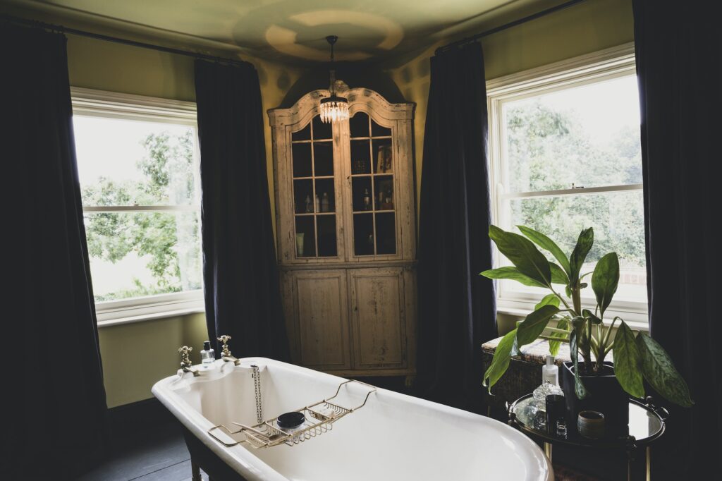 Interior view of bathroom with wooden corner cabinet between sash windows, roll top bath with brass